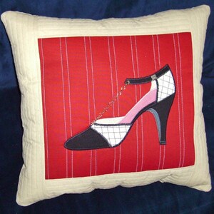 May include: Square decorative pillow with a cream-colored border and a red center featuring a high-heeled shoe design. The shoe is black, white, and pink with a gold chain detail. The red center has vertical white stripes.