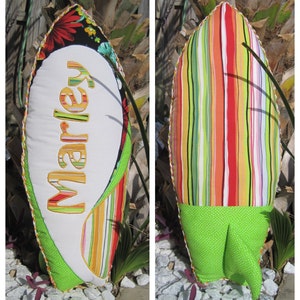 May include: A white and green surfboard-shaped cushion with the name "Marley" embroidered on the front. The back of the cushion is made of colourful striped fabric.