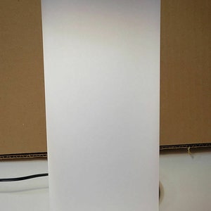 May include: A cylindrical white lamp with a frosted finish, illuminated from within. The lamp has a black cord and a simple, modern design, suitable for ambient lighting. The lamp is approximately 12 inches tall.