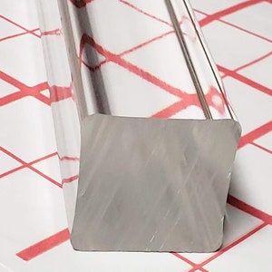 May include: A silver square bar with rounded edges. The bar is lying on a white surface with a red grid pattern.