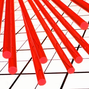 May include: Bright red cylindrical rods with rounded ends are arranged diagonally across a white grid background. The rods appear to be made of a translucent material, with visible internal patterns. The ends of the rods are a solid, opaque red.