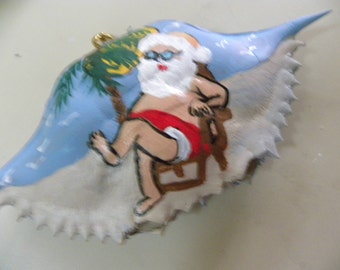 Santa Claus in bathing suit relaxing on beach painted on crab shell ornament. Packaged in a plastic container with coordinated shred