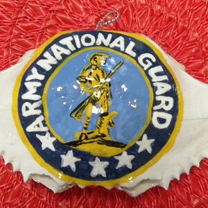 May include: A white crab shell ornament with a blue and yellow painted Army National Guard logo. The logo features a soldier holding a rifle and three stars.