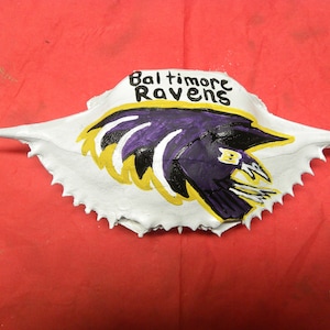 May include: A white crab shell painted with the Baltimore Ravens logo. The logo features a purple raven with yellow accents and the words "Baltimore Ravens" written above it.