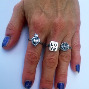 May include: Four silver rings are displayed on a hand with blue nail polish. The rings feature various designs: a heart with a crown, the word "LOVE", and a bird. The rings are made of a shiny metal.