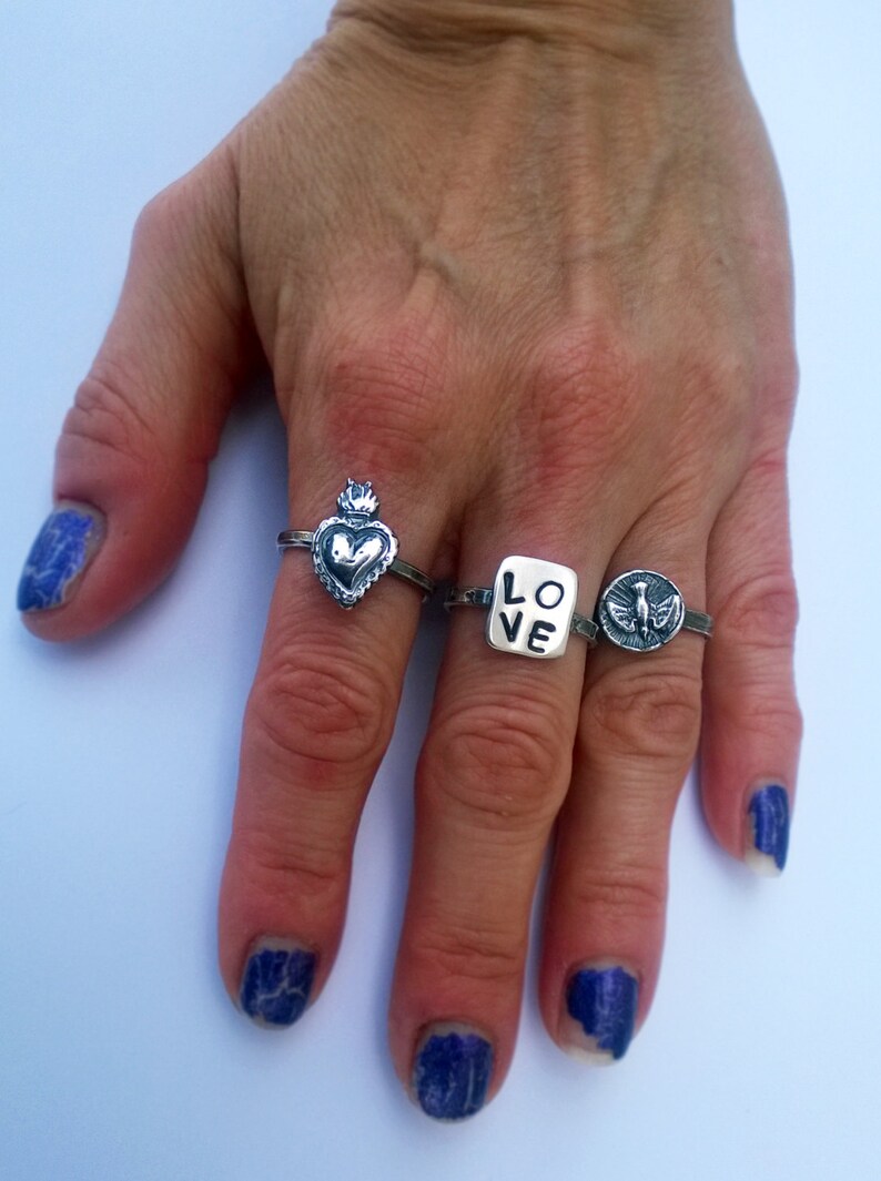 May include: Four silver rings are displayed on a hand with blue nail polish. The rings feature various designs: a heart with a crown, the word "LOVE", and a bird. The rings are made of a shiny metal.