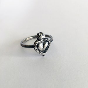 May include: A silver ring featuring a heart-shaped pendant with a crown design. The ring has a textured band and a detailed heart pendant. The metal has a slightly oxidized finish, giving it a vintage look. The ring is set against a white background.