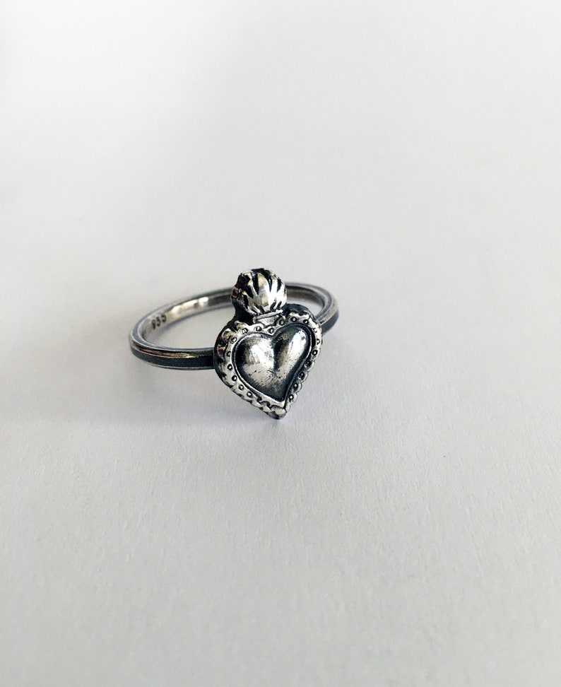 May include: A silver ring featuring a heart-shaped pendant with a crown design. The ring has a textured band and a detailed heart pendant. The metal has a slightly oxidized finish, giving it a vintage look. The ring is set against a white background.