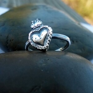 May include: A silver ring featuring a heart-shaped design with a crown on top. The heart has a textured surface and is surrounded by a decorative border. The ring is set on a dark, smooth stone.