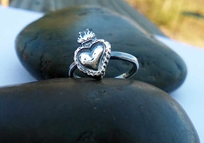 May include: A silver ring featuring a heart-shaped design with a crown on top. The heart has a textured surface and is surrounded by a decorative border. The ring is set on a dark, smooth stone.