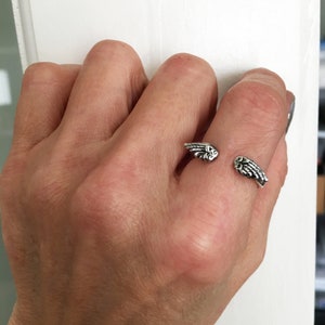 May include: A silver ring with a wing design. The ring is worn on the middle finger of a person's hand.