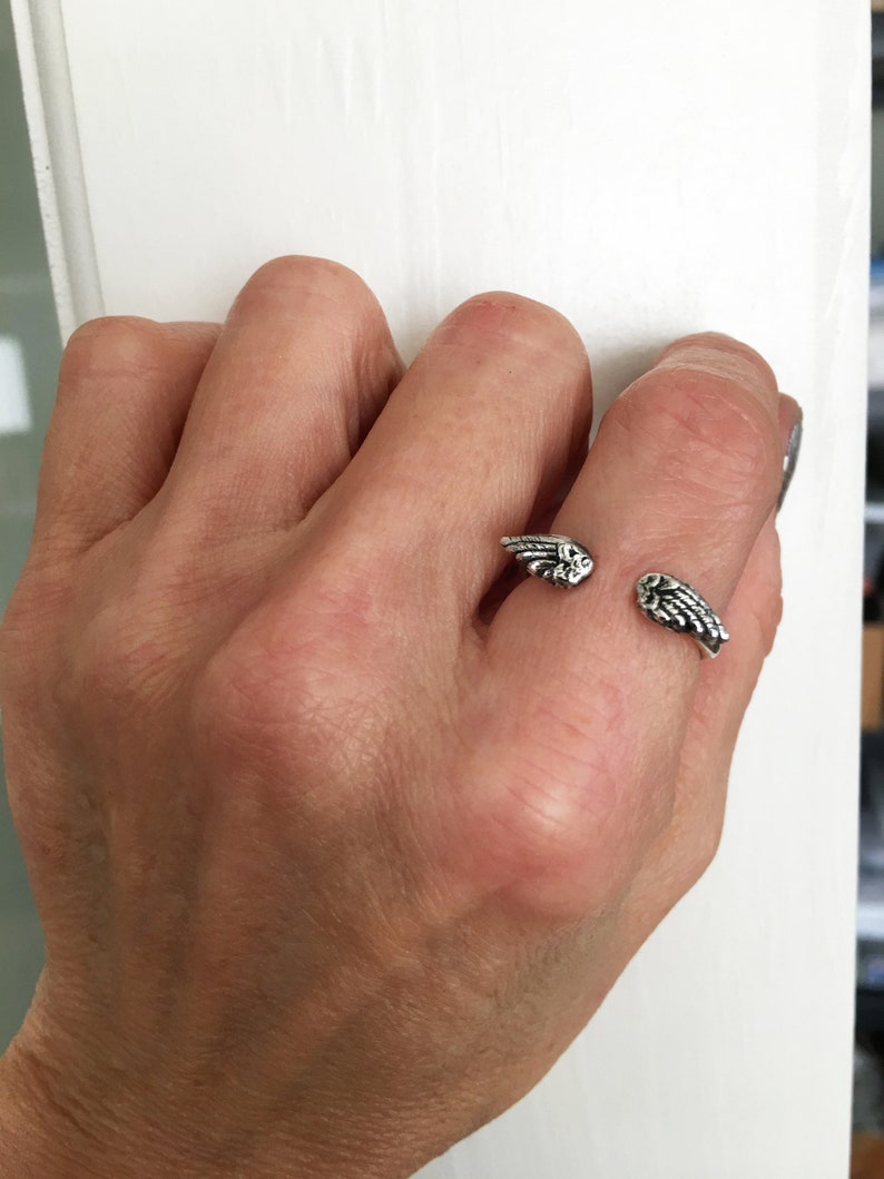 May include: A silver ring with a wing design. The ring is worn on the middle finger of a person's hand.