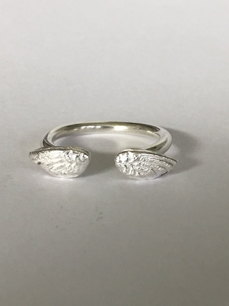 May include: A silver ring with two angel wings on either side. The wings are detailed and have a textured finish.