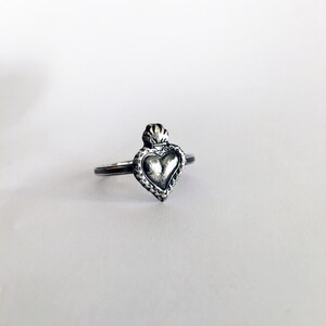 May include: A silver-toned ring featuring a heart-shaped pendant with a crown design. The heart has a textured surface and is surrounded by a decorative border. The ring is set against a white background.