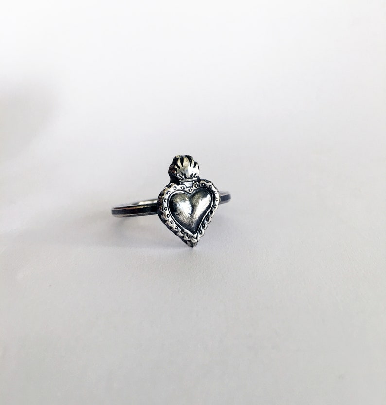 May include: A silver-toned ring featuring a heart-shaped pendant with a crown design. The heart has a textured surface and is surrounded by a decorative border. The ring is set against a white background.