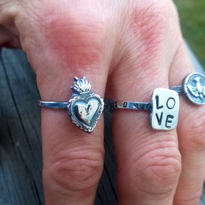 May include: A hand displays several silver rings. One ring features a heart design with a flame on top. Another ring has the word "LOVE" on a rectangular plate. A third ring has a bird emblem.