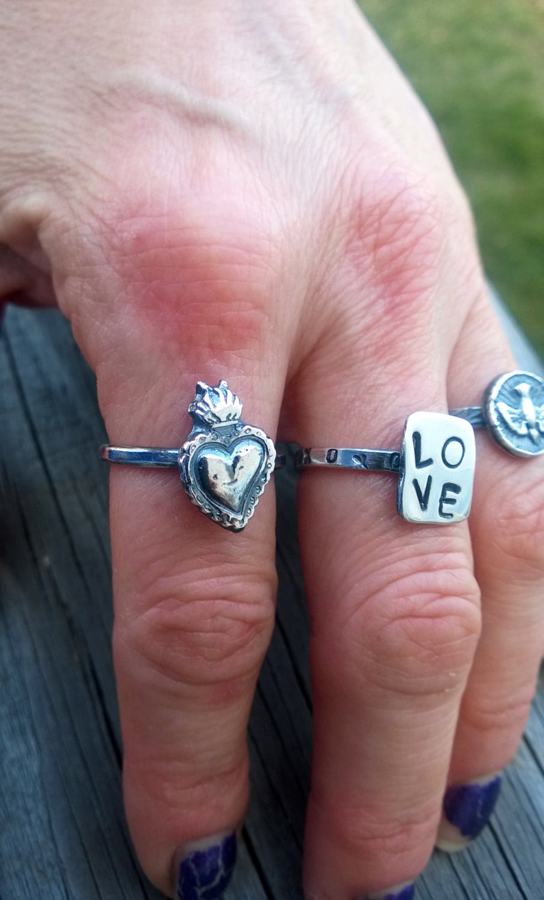 May include: A hand displays several silver rings. One ring features a heart design with a flame on top. Another ring has the word "LOVE" on a rectangular plate. A third ring has a bird emblem.