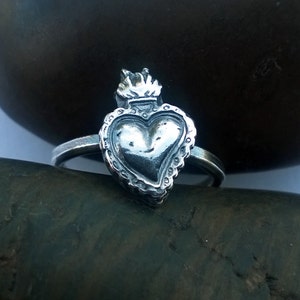 May include: A silver ring featuring a detailed heart design with a crown and flame accents. The heart is the focal point, surrounded by a decorative border. The ring is set against a dark, textured background.
