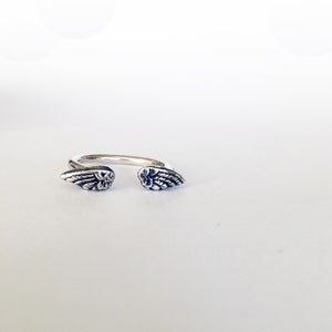 May include: A silver ring with two angel wings on either side. The wings are detailed and have a textured finish.