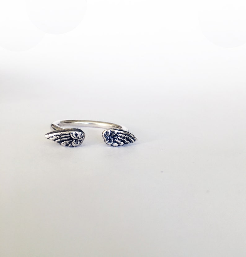 May include: A silver ring with two angel wings on either side. The wings are detailed and have a textured finish.