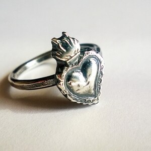 May include: A silver ring featuring a heart-shaped design with a crown on top. The ring has a detailed, ornate border around the heart. The ring is set against a white background.