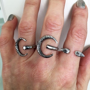 May include: Three silver rings on a hand. The rings are a crescent moon with a star, a crescent moon with a detailed design, and a pair of angel wings.
