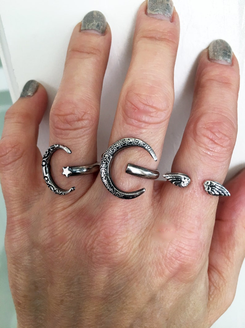 May include: Three silver rings on a hand. The rings are a crescent moon with a star, a crescent moon with a detailed design, and a pair of angel wings.