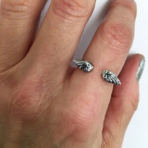 May include: A silver ring with two wings on the top. The ring is on a person's finger.