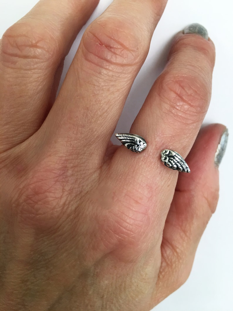 May include: A silver ring with two wings on the top. The ring is on a person's finger.