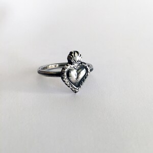 May include: A silver ring featuring a heart-shaped pendant with a detailed, textured design. The heart is topped with a flame-like element. The ring has a thin band and is set against a white background.