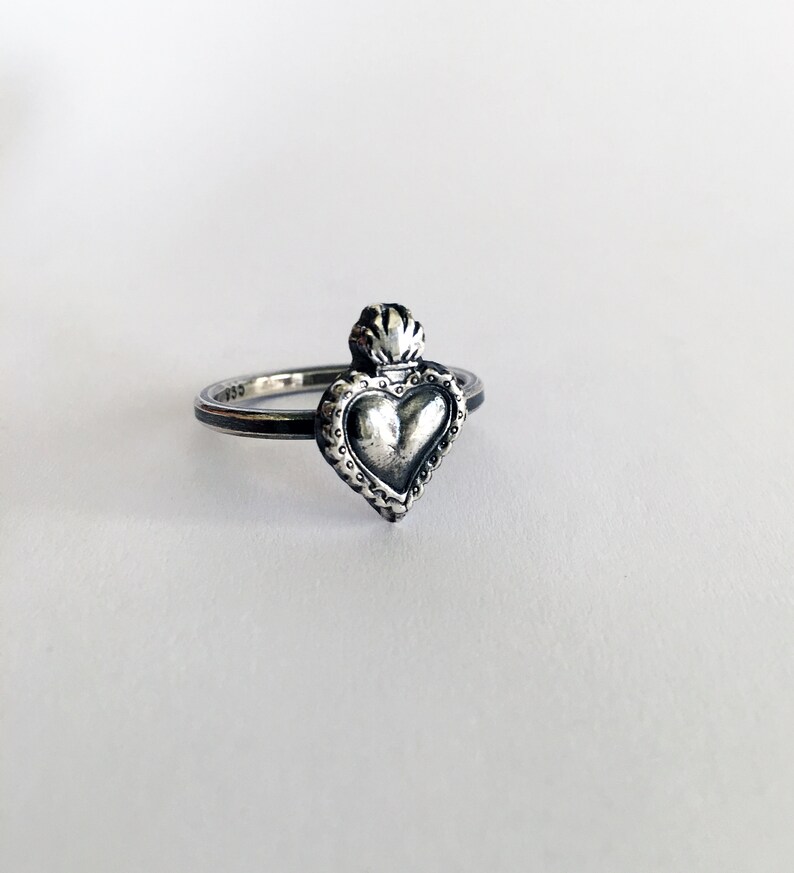 May include: A silver ring featuring a heart-shaped pendant with a detailed, textured design. The heart is topped with a flame-like element. The ring has a thin band and is set against a white background.
