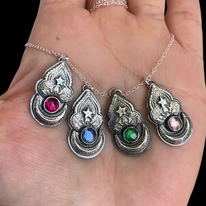 May include: Four silver pendant necklaces with different colored gemstones: red, blue, green, and pink. Each pendant has a star and ornate design. The necklaces are on a silver chain.