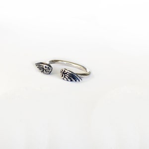 May include: A silver ring with two angel wings on either side. The wings are detailed and have a textured finish.