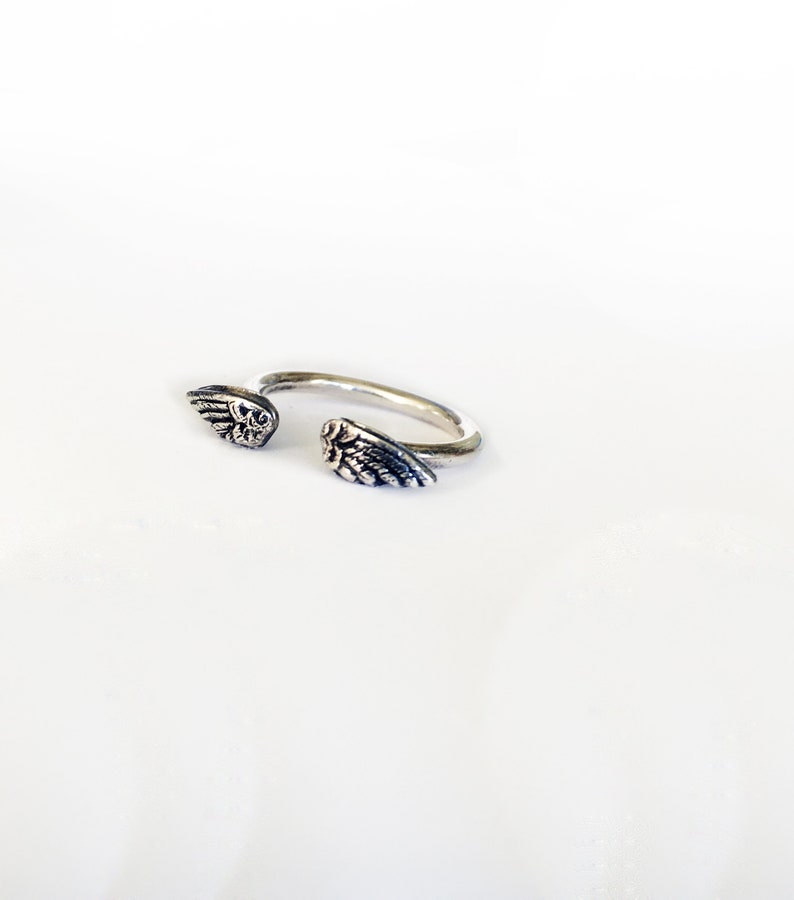 May include: A silver ring with two angel wings on either side. The wings are detailed and have a textured finish.