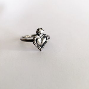 May include: A silver ring featuring a heart-shaped pendant with a crown-like detail at the top. The heart has a textured surface and is surrounded by a decorative border. The ring is set against a white background.