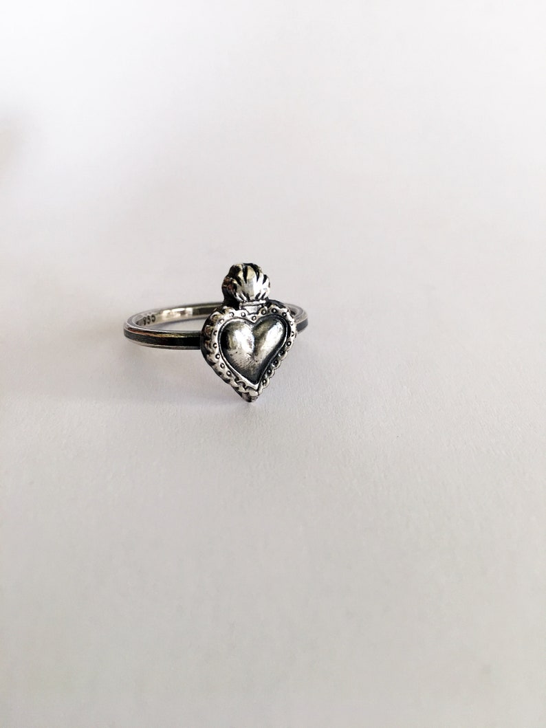 May include: A silver ring featuring a heart-shaped pendant with a crown-like detail at the top. The heart has a textured surface and is surrounded by a decorative border. The ring is set against a white background.