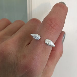 May include: A silver ring with two wings on the band. The ring is on a person's finger.