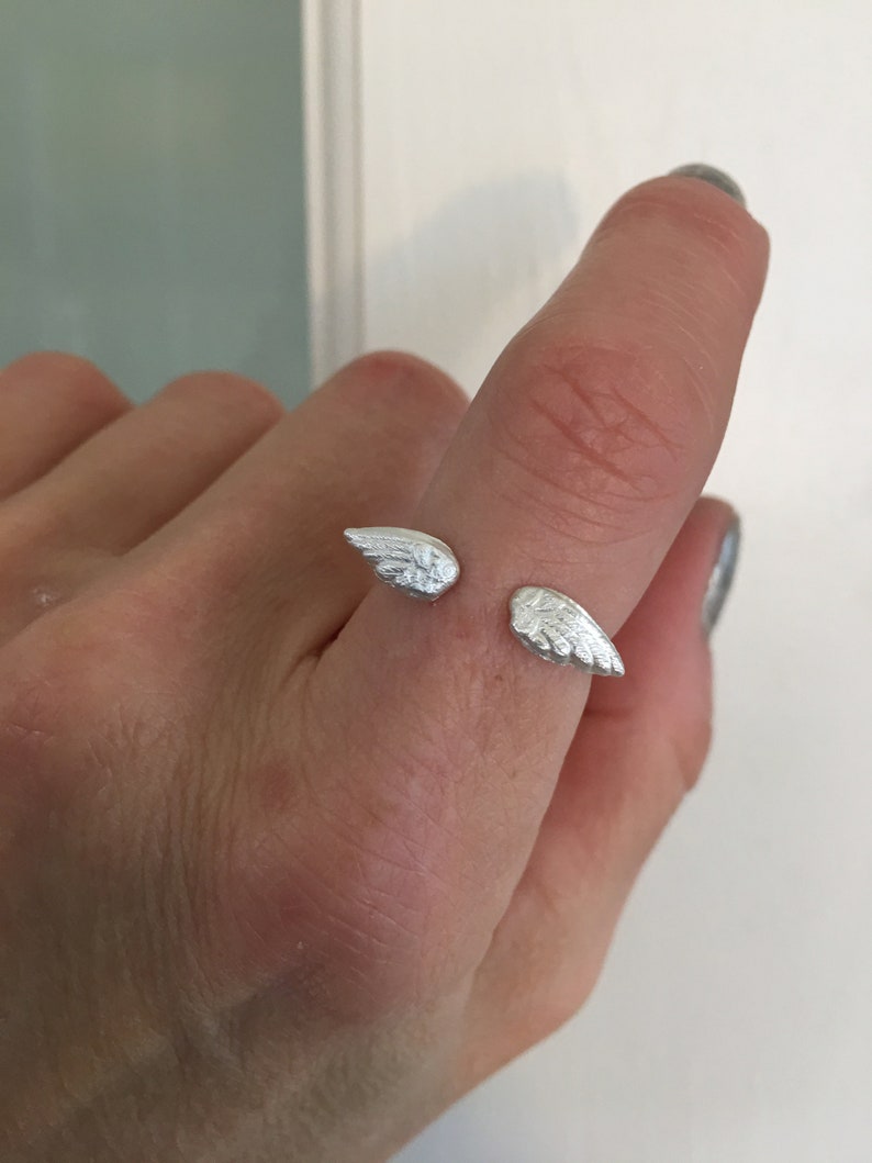 May include: A silver ring with two wings on the band. The ring is on a person's finger.