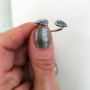 May include: A silver ring with two angel wings on either side. The ring is open and is being held by a finger with silver nail polish.