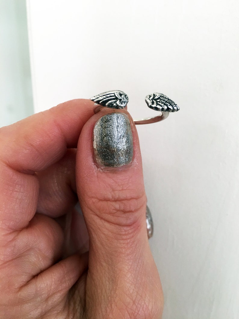 May include: A silver ring with two angel wings on either side. The ring is open and is being held by a finger with silver nail polish.