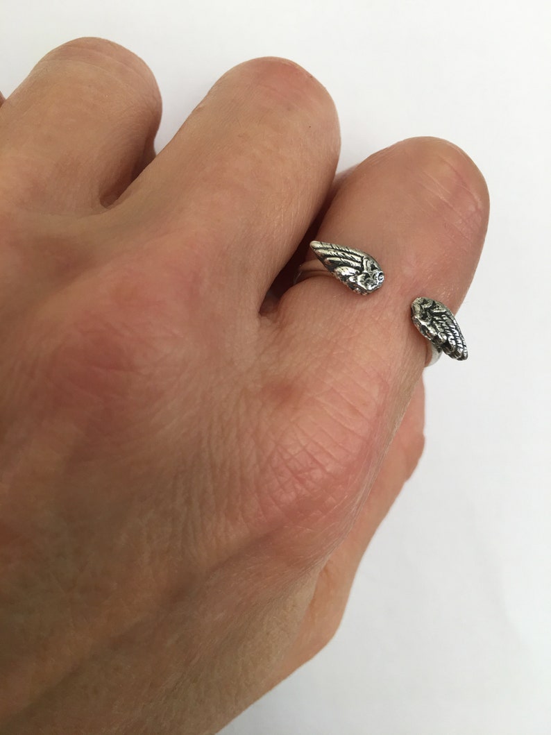 May include: A silver ring with two angel wings on the band. The wings are detailed and have a textured finish.