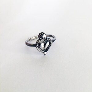 May include: A silver ring featuring a heart-shaped pendant with a crown on top. The heart has a textured surface and is surrounded by a beaded border. The ring is set against a white background.