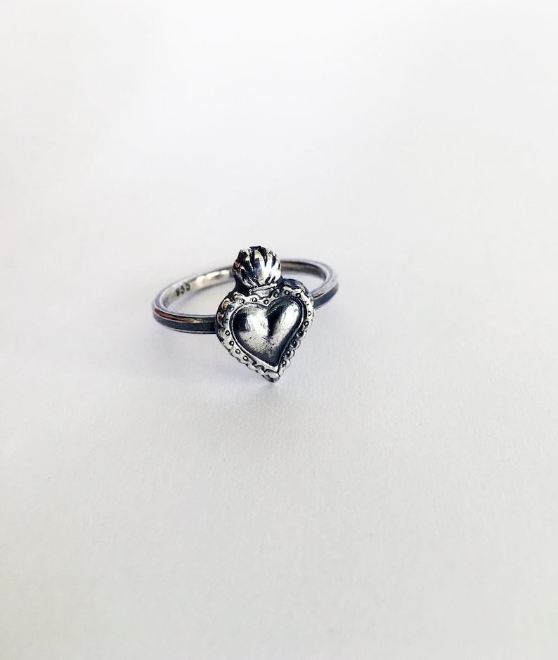 May include: A silver ring featuring a heart-shaped pendant with a crown on top. The heart has a textured surface and is surrounded by a beaded border. The ring is set against a white background.