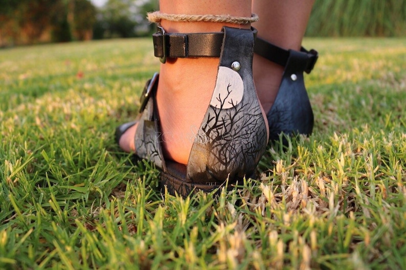May include: Black leather sandals with a painted design of a tree and moon on the back strap. The sandals have a buckle closure and a silver rivet.
