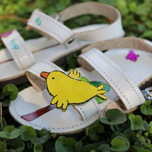 May include: White leather sandals with a yellow bird design and a purple flower design. The sandals have adjustable straps and a buckle fastening.