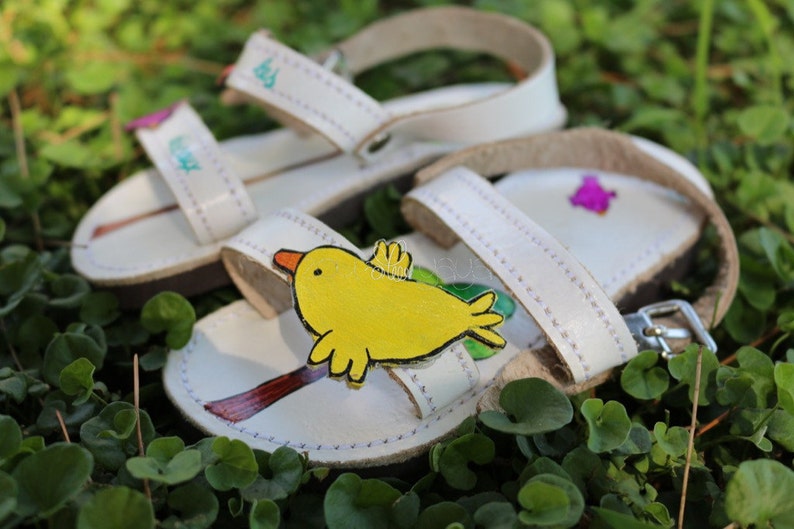 May include: White leather sandals with a yellow bird design and a purple flower design. The sandals have adjustable straps and a buckle fastening.