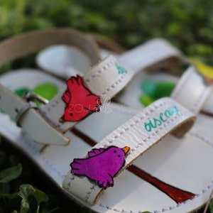 May include: White leather sandals with colourful bird appliques and the word "oiscay" written on the strap. The sandals have a simple design and are perfect for summer.