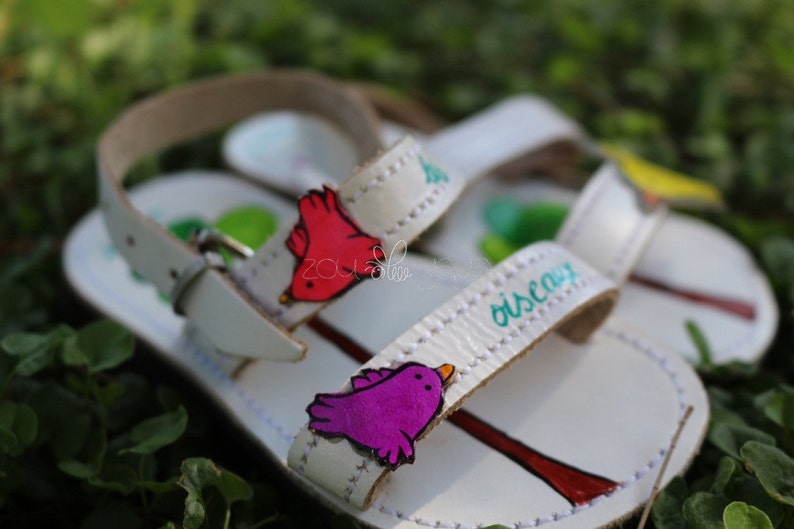 May include: White leather sandals with colourful bird appliques and the word "oiscay" written on the strap. The sandals have a simple design and are perfect for summer.