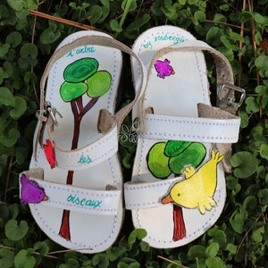 May include: White leather sandals with hand-painted designs. One sandal features a tree with green leaves and the text "l'arbre les oiseaux". The other sandal features a yellow bird and a tree with green leaves.