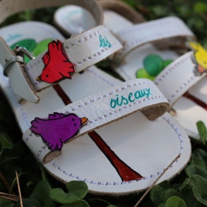May include: White leather sandals with colourful bird appliques and the text "les oiseaux" on the strap.